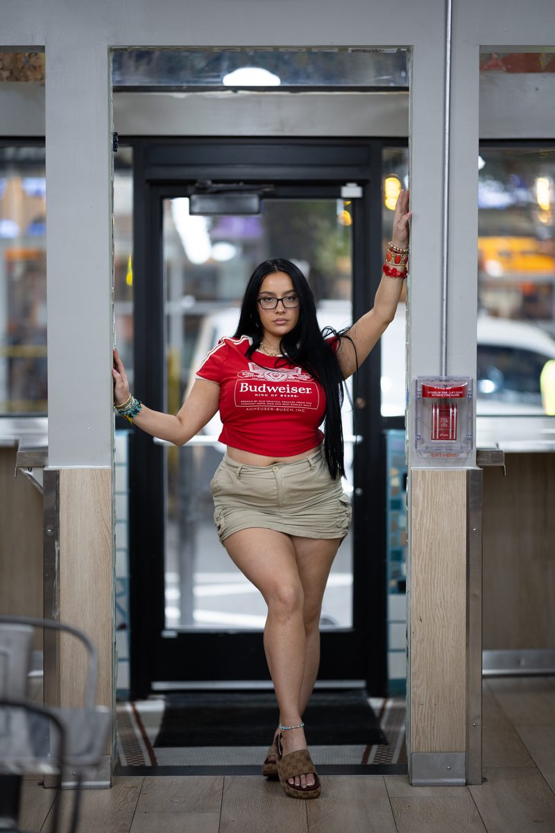 A woman in a red Budweiser crop top and khaki skirt poses in the doorway of a Jim's Steaks Philly cheesesteak shop on South Street with her arms posed on the frame of the doorway.