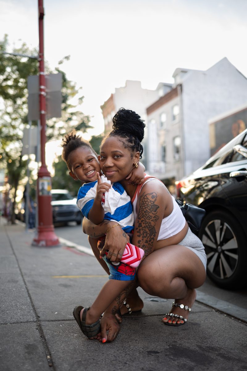 A mother with tattooed arms crouches to hold her young son, who has a happy smile and points up at the sky on a South Street sidewalk.