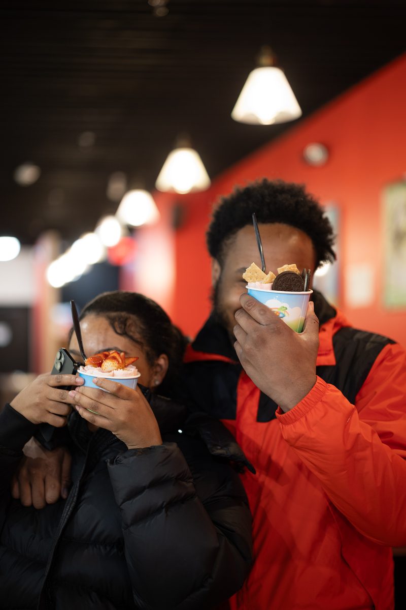 Two friends in puffer jackets hold cups of ice cream in front of their faces inside a Frozen Rolled Ice Cream shop on South Street.