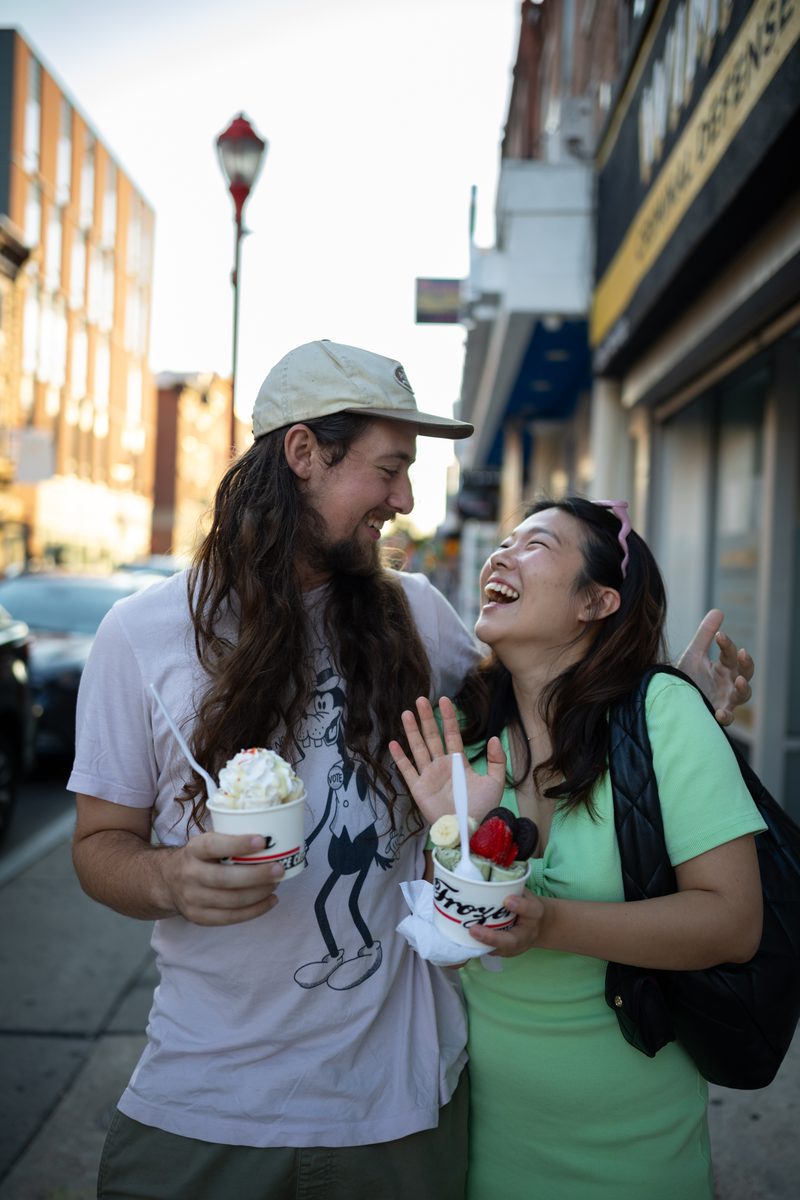A couple laughs together holding cups of Ice Cream from Frozen Rolled Ice on South Street, he with long hair and a cap. She is wearing a pastel green shirt.