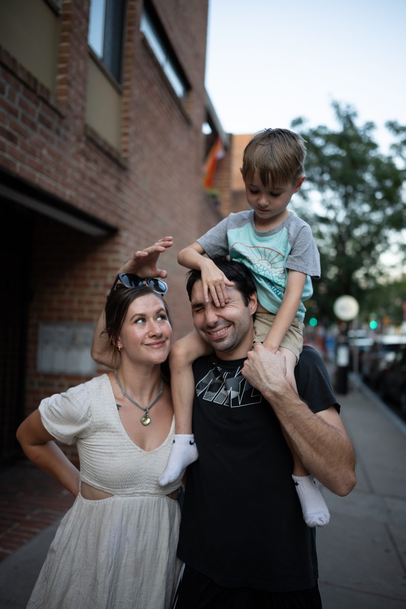 A woman in a white dress leans into a man carrying their young son on his shoulders on a South Street sidewalk. The young boy is playfully trying to poke his father's eye, while the father laughs, and the woman looks up at the son smiling.
