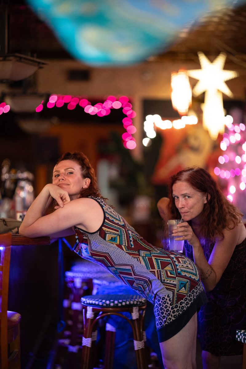 Two women rest their chins on their hands at Copa Banana bar on South Street under pink string lights, one in a colorful patterned dress.