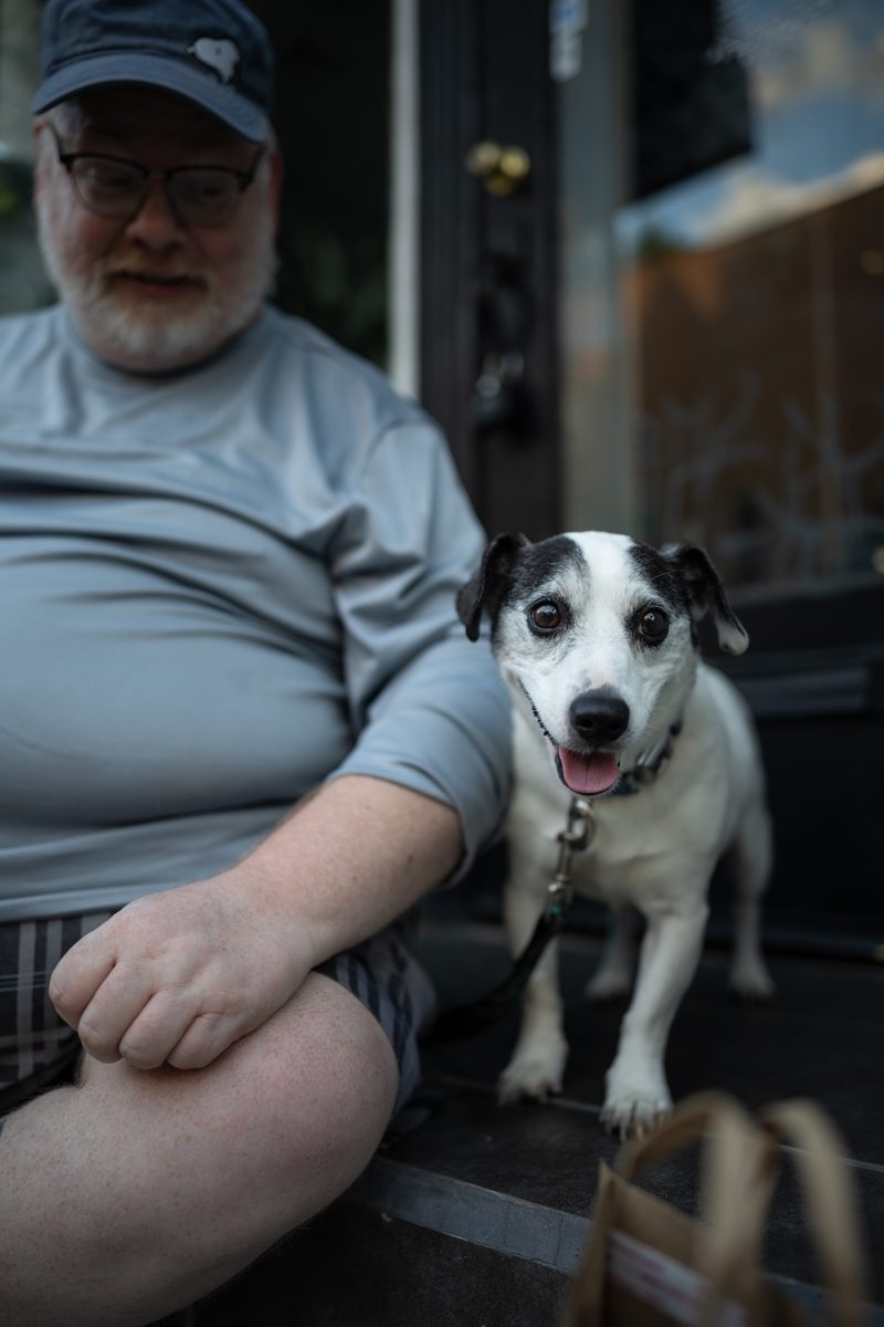 A man in a gray t-shirt and cap sits on a step with his black-and-white dog, who looks straight at the camera.