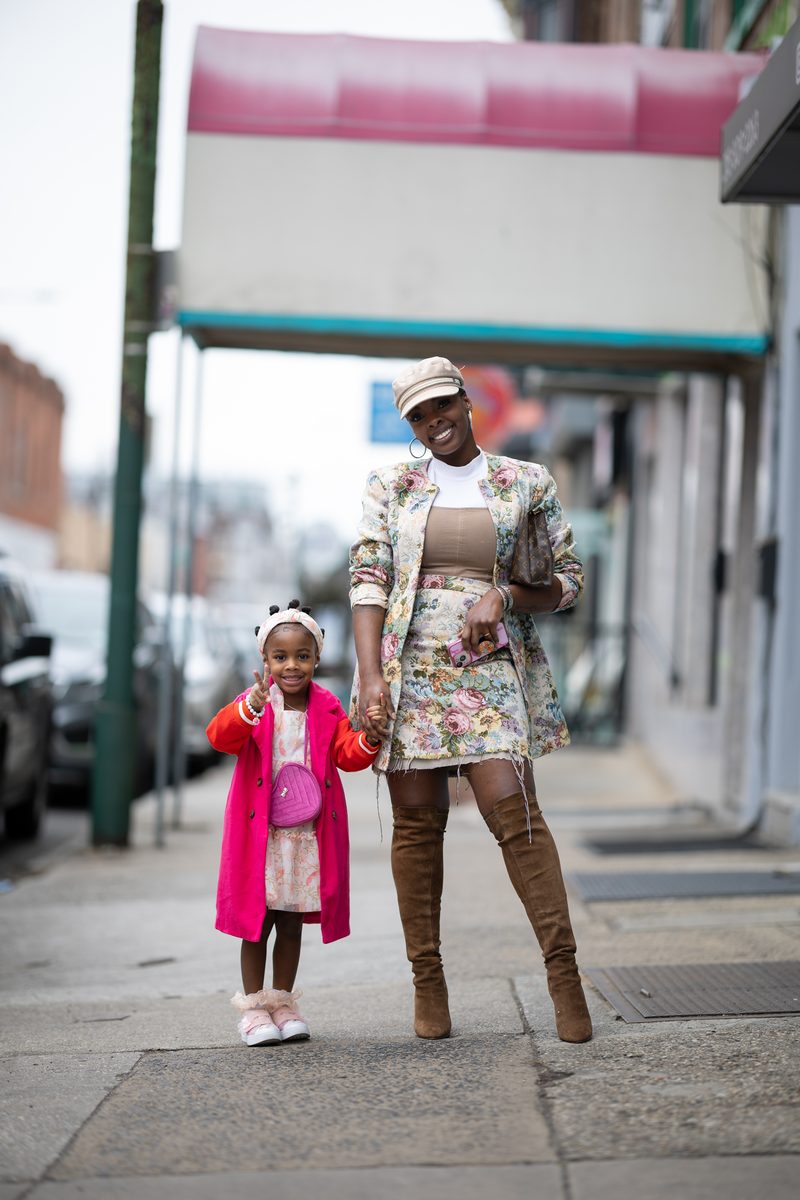 A mother in a floral suit and tall suede boots holds hands with her young daughter in a pink coat on South Street. They are both smiling, with the daughter posing by making a peace sign with her hand.
