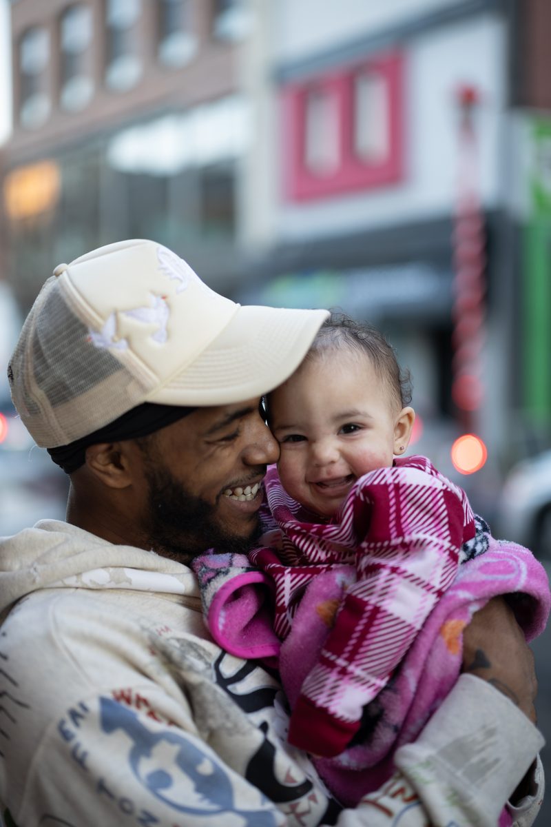 A father in a white trucker hat laughs with his baby daughter, wrapped in a pink plaid blanket, on South Street.