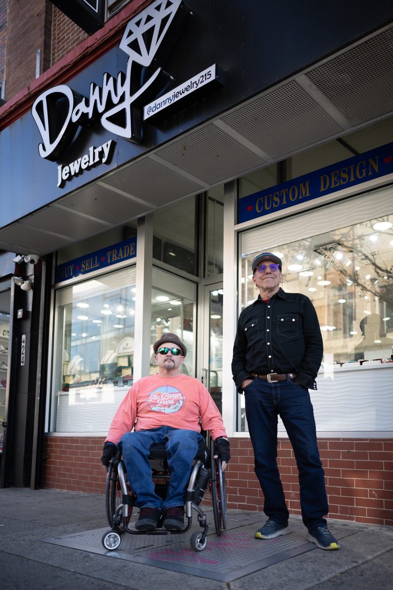 Two men pose outside Danny Jewelry on South Street, one in a wheelchair in a coral sweatshirt, the other in black.