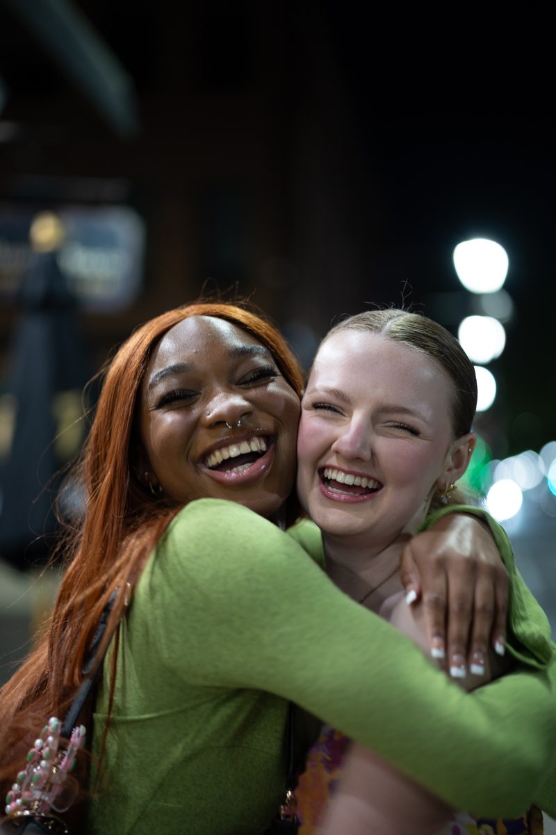 Two friends hug and laugh on South Street at night, one with long red hair, the other in a green top.