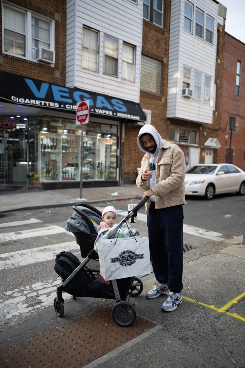 A father in a hoodie pushes his baby in a stroller across a South Street crosswalk in front of Vegas smoke shop.