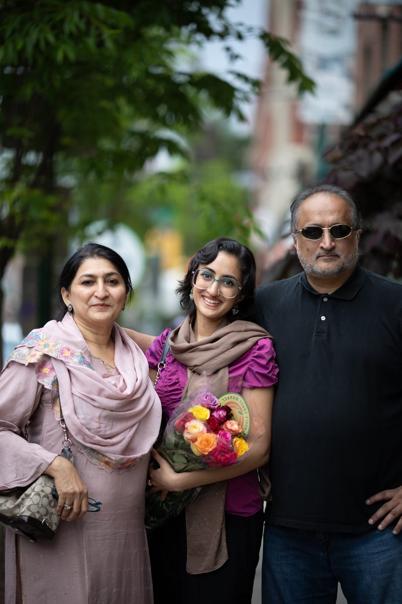 A family of three, the mother in a pink shawl and the daughter holding a bouquet of flowers, stands together on South Street.