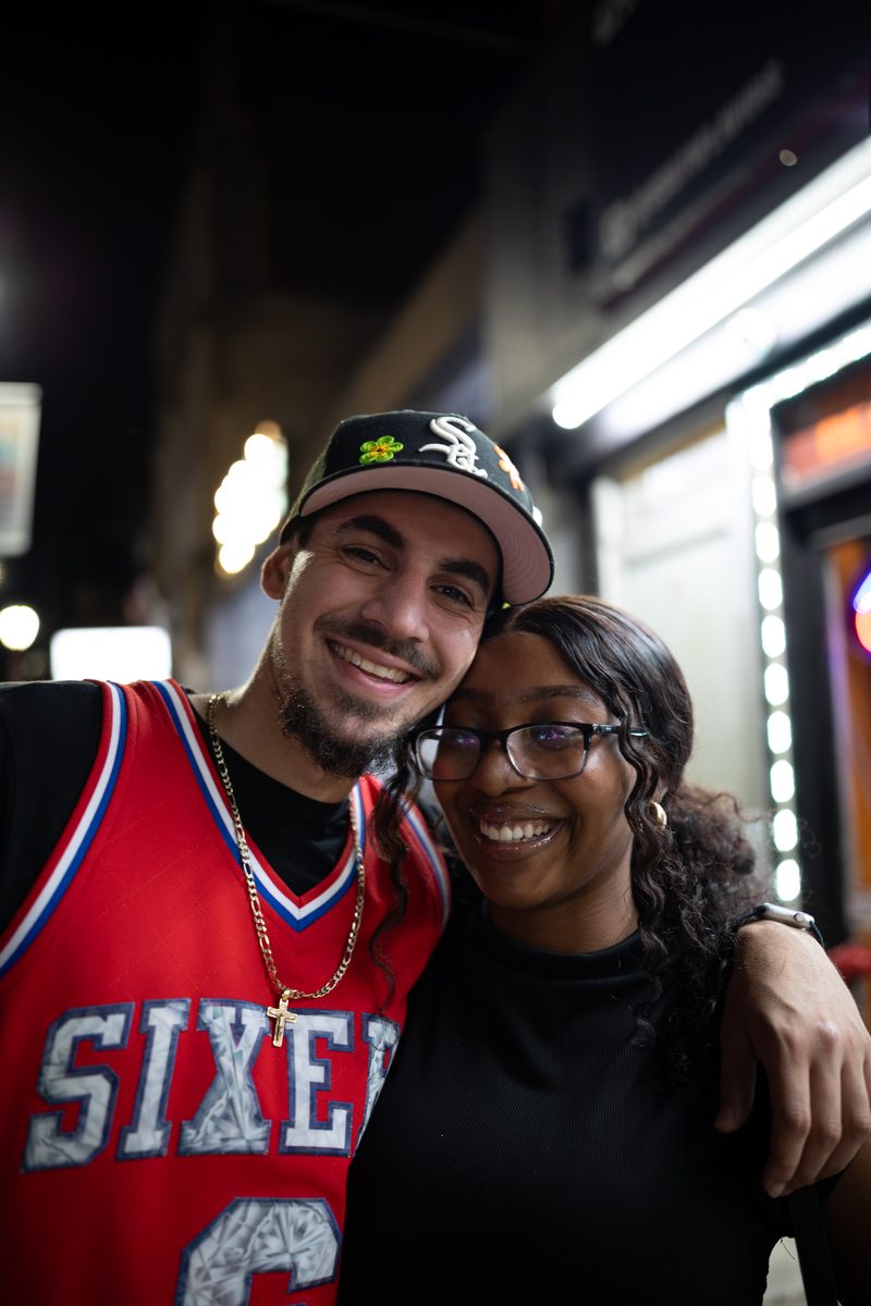 A couple smiles cheek to cheek on South Street at night, he in a red Sixers jersey and White Sox cap.