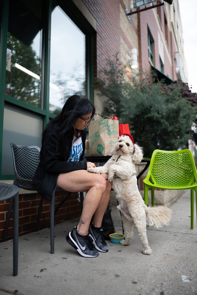 A woman at a sidewalk café table by Whole Foods supermarket on South Street holds the paws of her cream-colored poodle mix, who stands on hind legs.