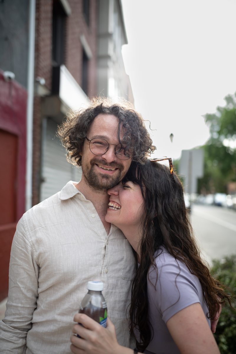 A curly-haired man in glasses laughs as his partner presses her forehead against his neck, smiling, on a South Street sidewalk.