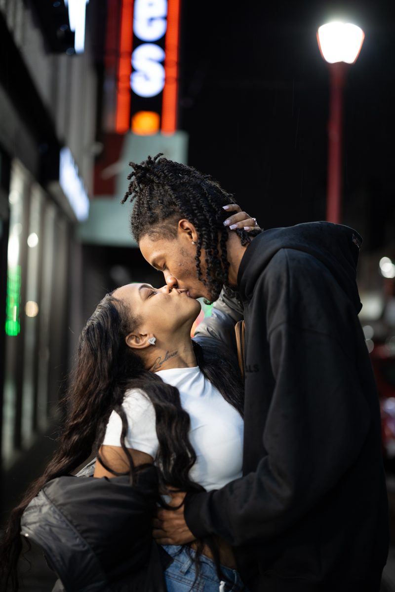 A couple shares a passionate kiss under a streetlight on a sidewalk in Philadelphia, the neon sign for Snipes flagship store on South Street in orange behind them.