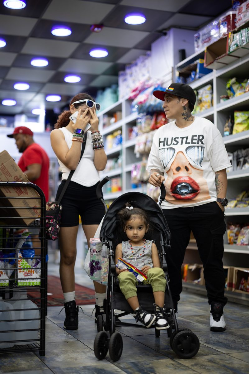 A family of three walks through a South Street corner store, the toddler in a stroller looking straight at the camera and one of the parents wearing a "Hustler" T-shirt.